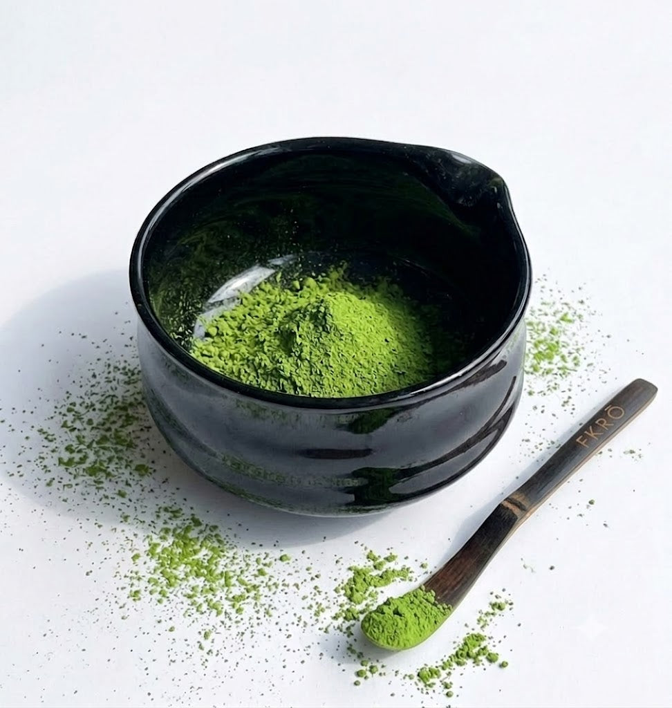 Hand holding a FKRŌ bamboo chasen whisking thick vivid green matcha in a black chawan bowl with gold teaware arranged in the background