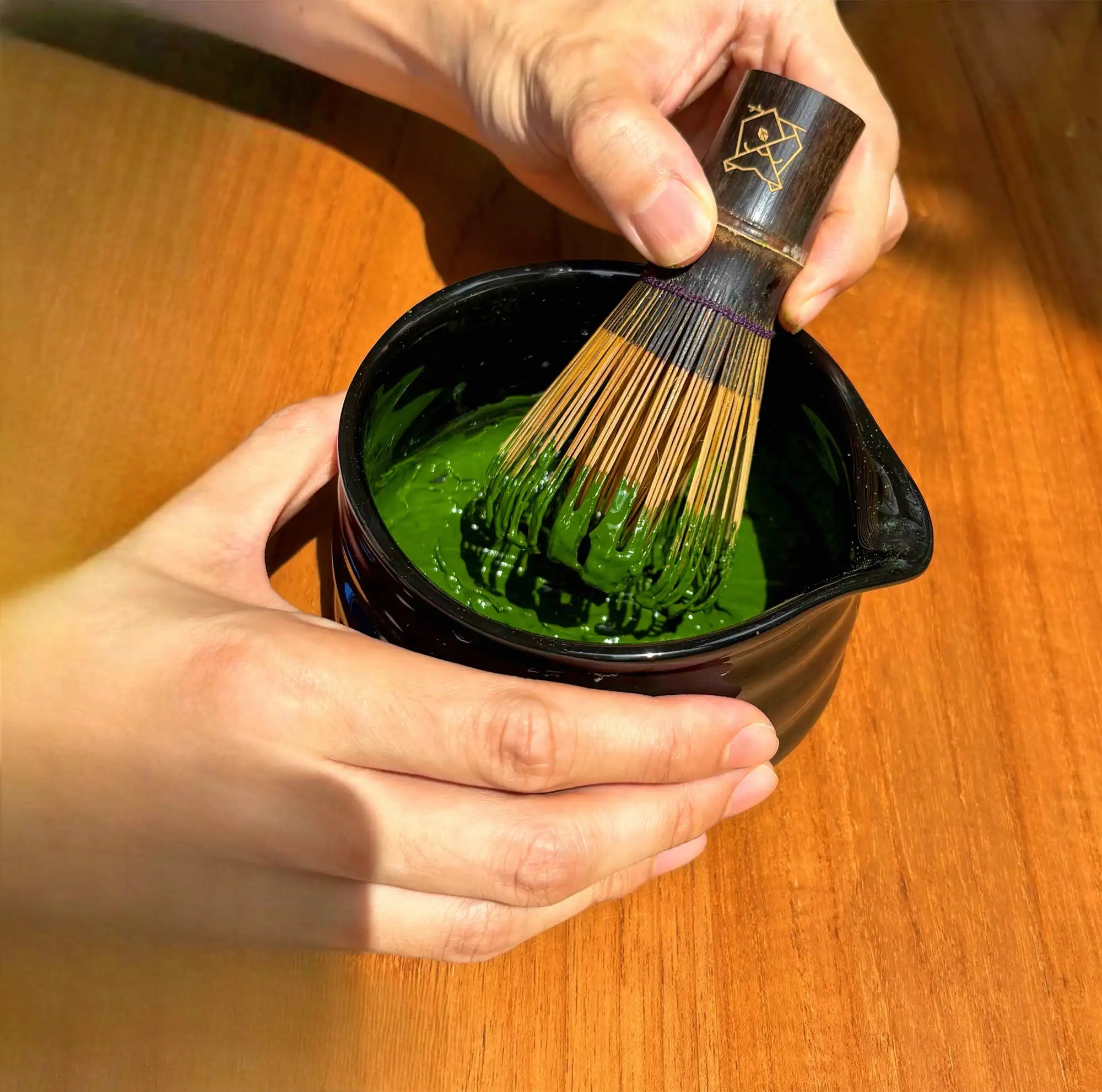 Black ceramic matcha bowl with green tea mixture and a bamboo whisk being used to stir it.