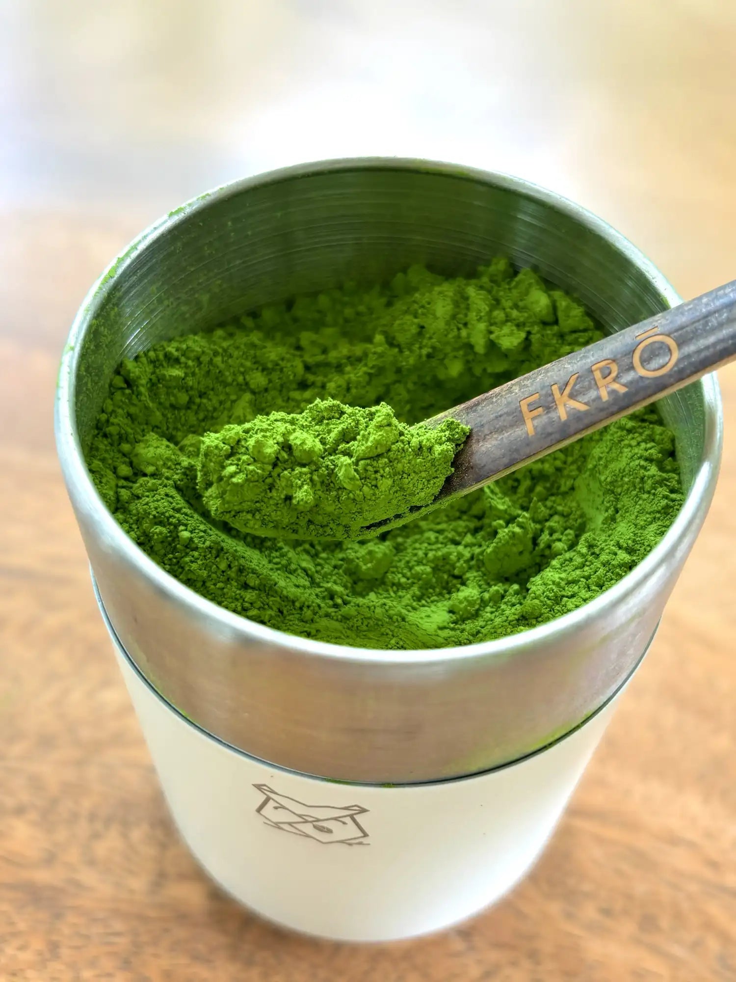 A metallic container filled with vibrant green matcha powder, featuring a wooden spoon branded ’fkrö’ resting inside.