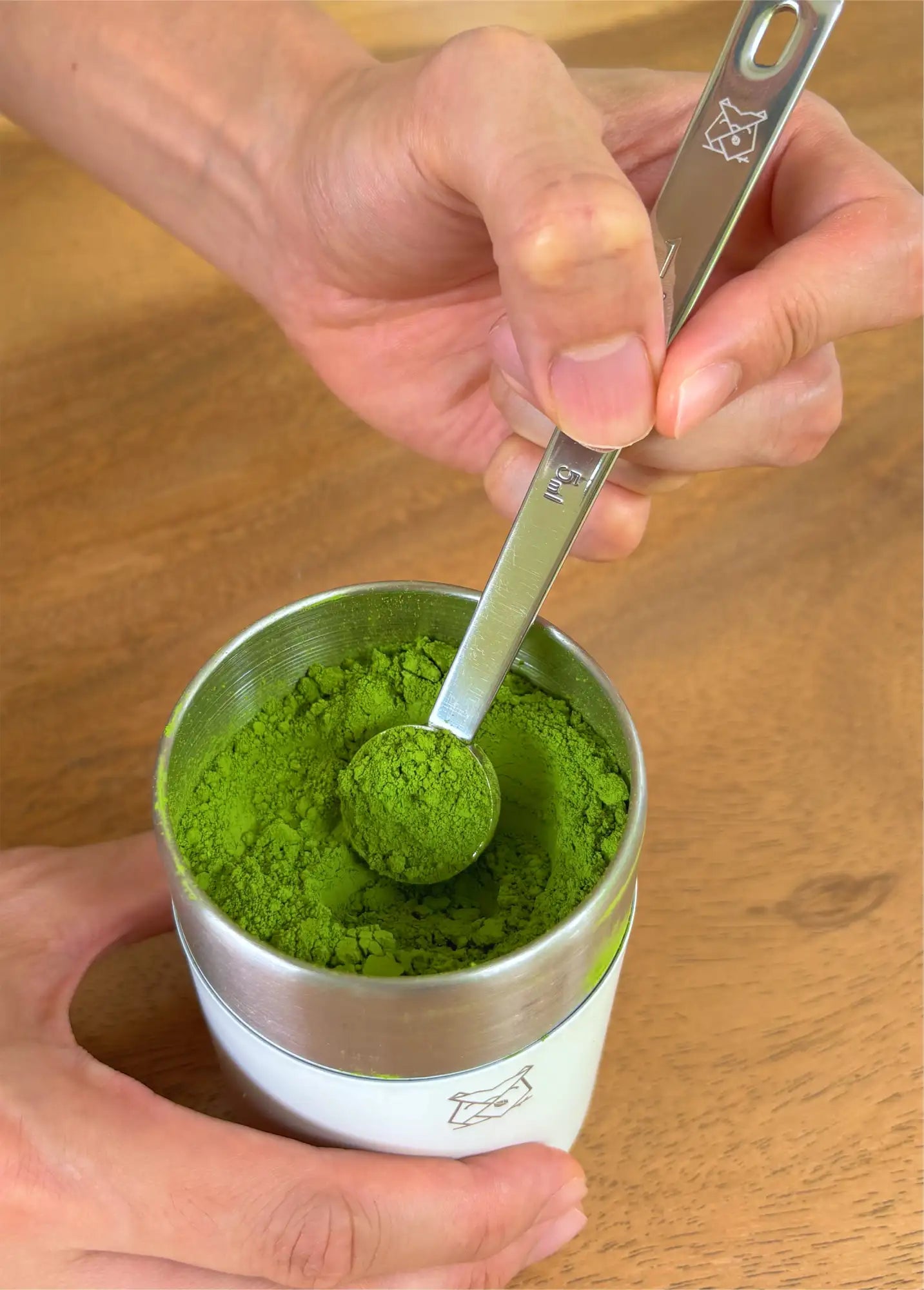 A stainless steel measuring spoon filled with vibrant green matcha powder, held above a white and silver ceramic container.