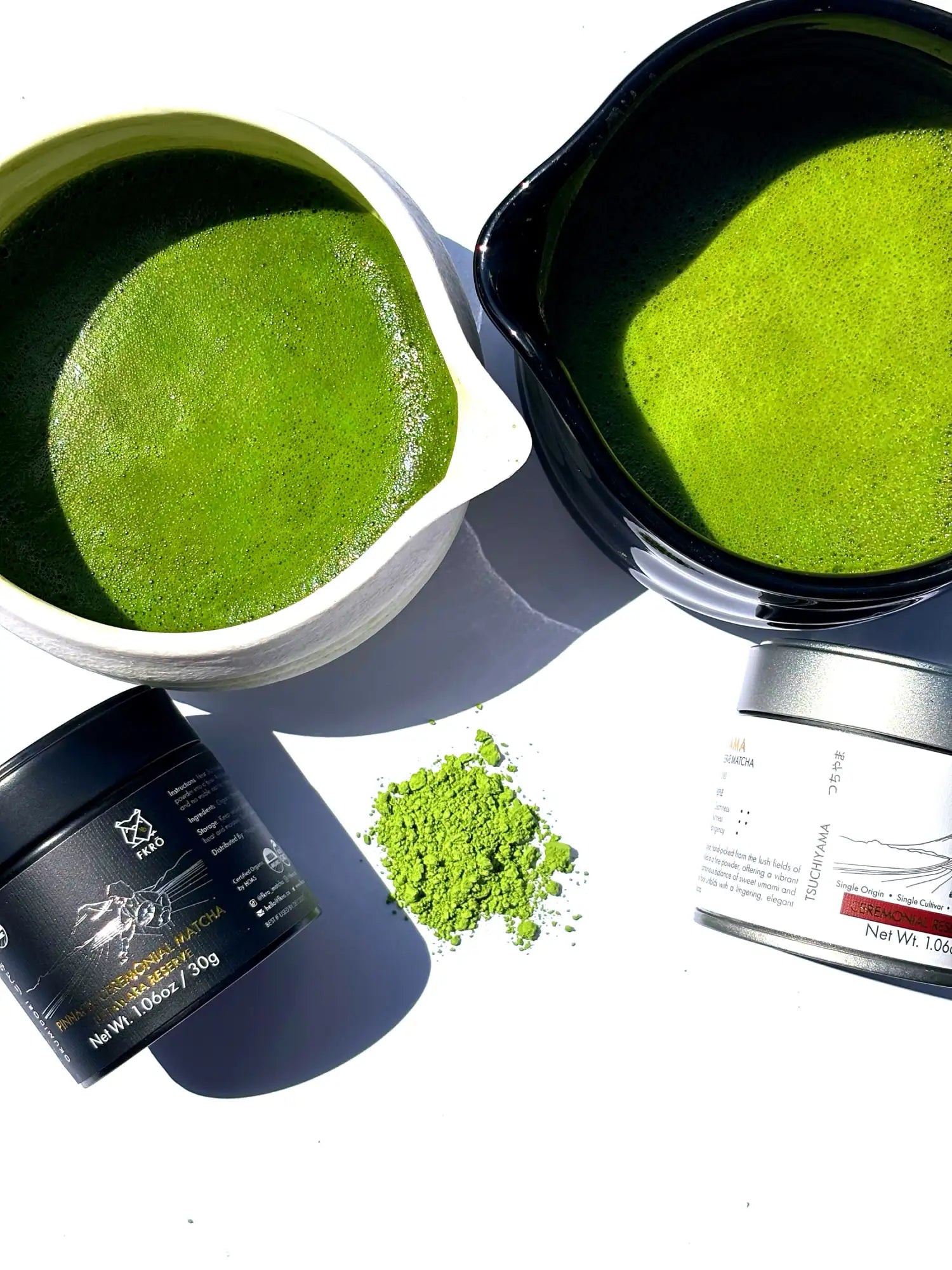 Two ceramic bowls filled with vibrant green matcha tea, accompanied by a black container of powdered matcha and a silver tin labeled ’matcha.’