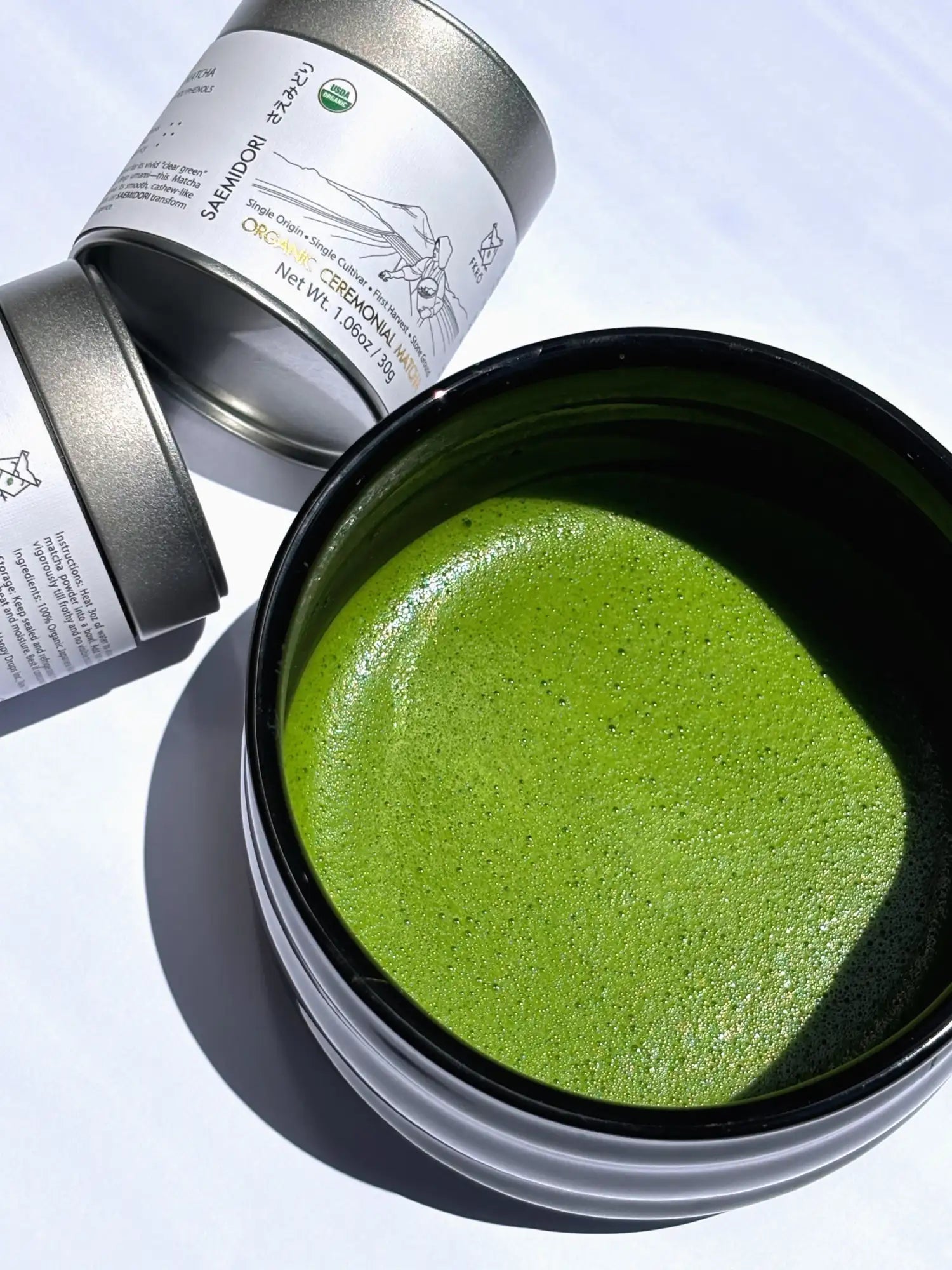 A vibrant, frothy green matcha tea served in a sleek black ceramic bowl beside its metallic tin container.