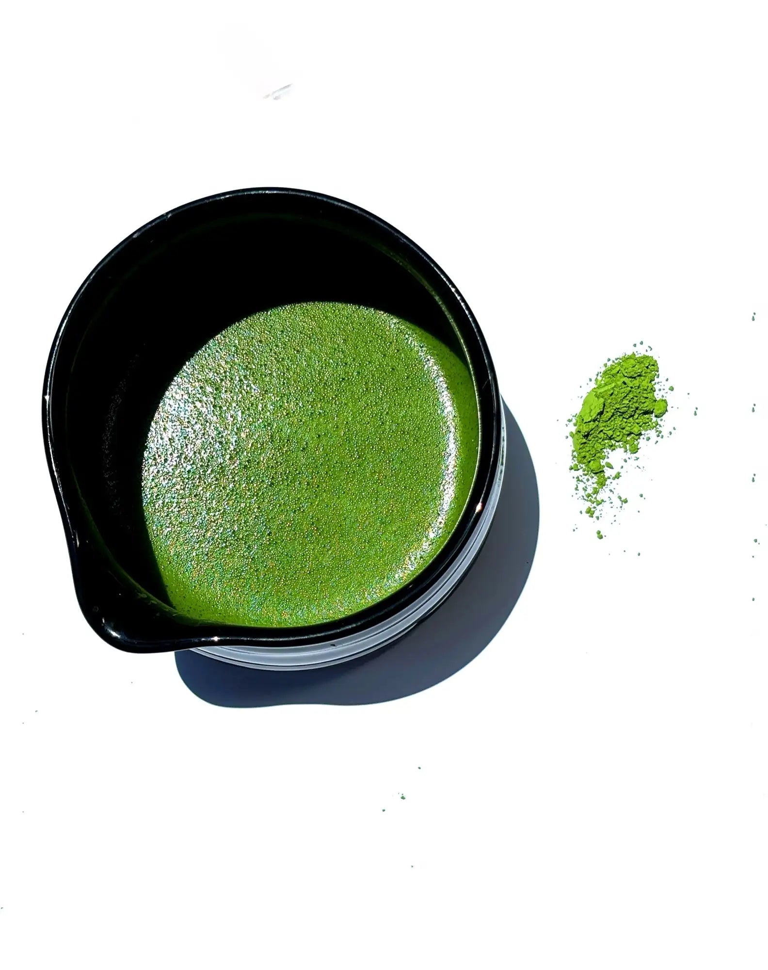 A vibrant green matcha latte in a sleek black ceramic bowl, with a small pile of loose matcha powder beside it.